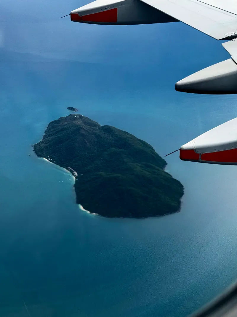 Forested island surrounded by deep blue sea seen past an airliner wing