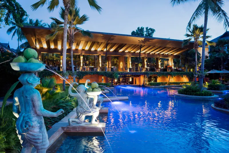 Lit pool terrace and open-sided restaurant at dusk at Anantara Bophut Koh Samui, with fountain statues