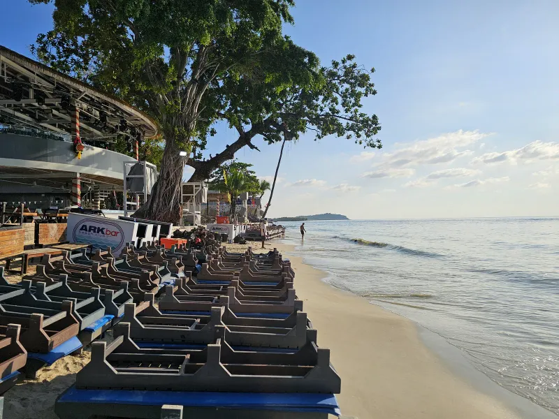 Sandy shoreline at Chaweng Beach with beach club loungers stacked beside a large tree
