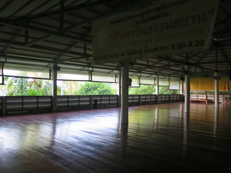 Interior of the open-sided meditation hall with a notice board and polished teak floor