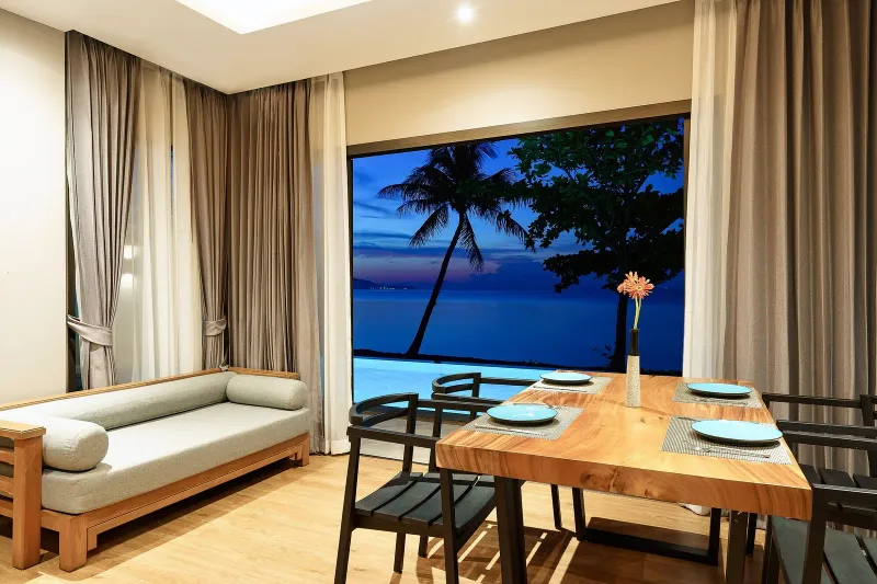Villa living and dining room at dusk with a wide window onto a pool and the sea