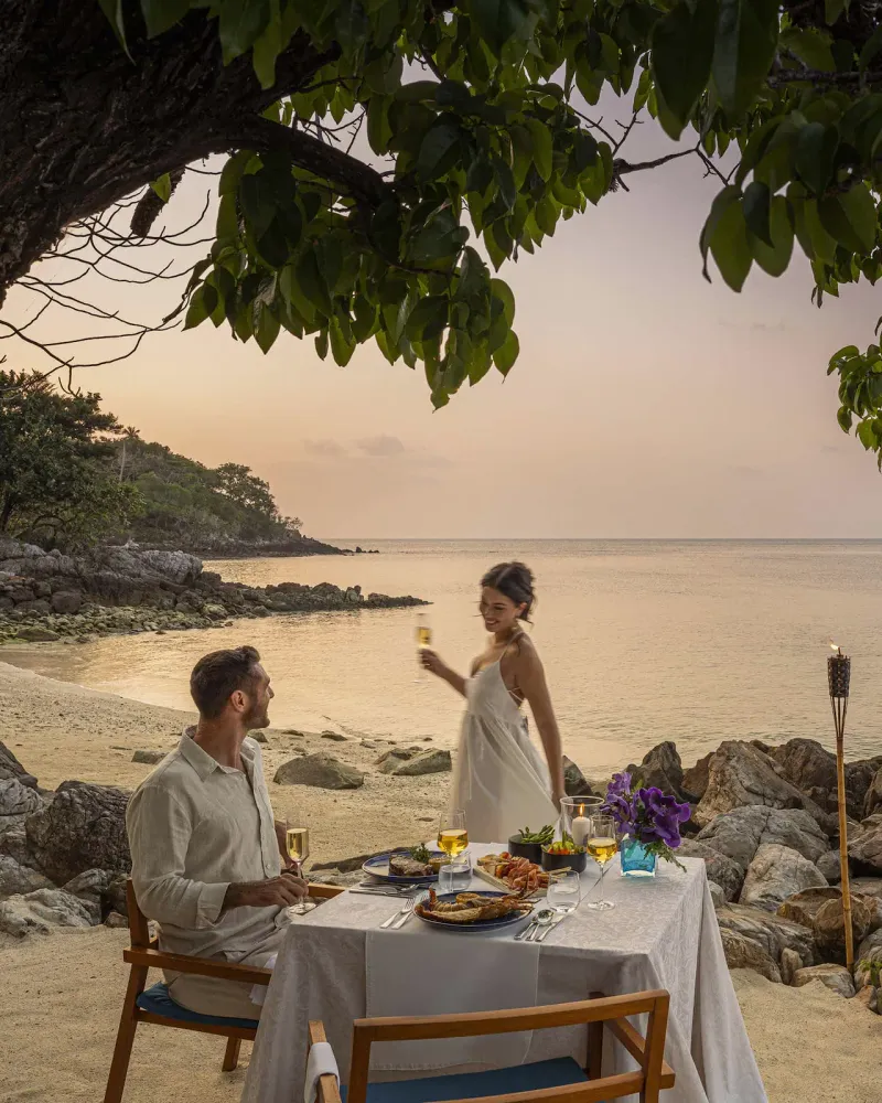 Couple at a private table set on the sand at dusk with a torch-lit rocky shore at Four Seasons Resort Koh Samui