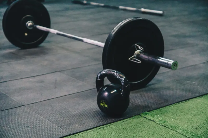 Loaded barbell and kettlebell resting on a rubber gym floor beside artificial turf strips