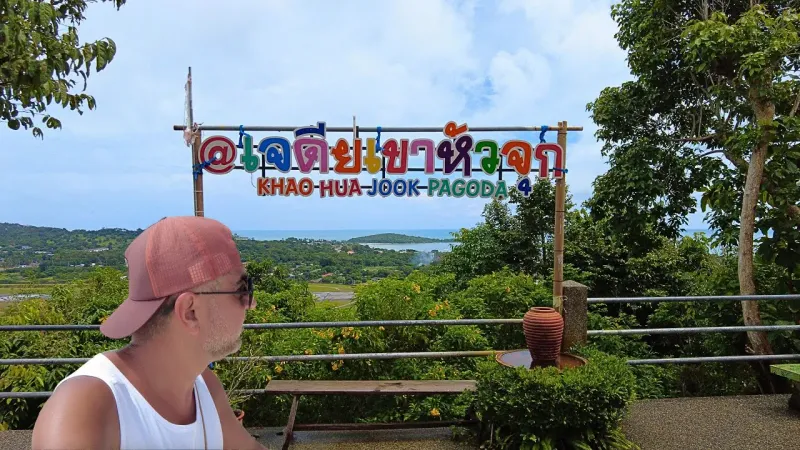 A visitor beside the Khao Hua Jook Pagoda sign, with the coastline and sea visible below