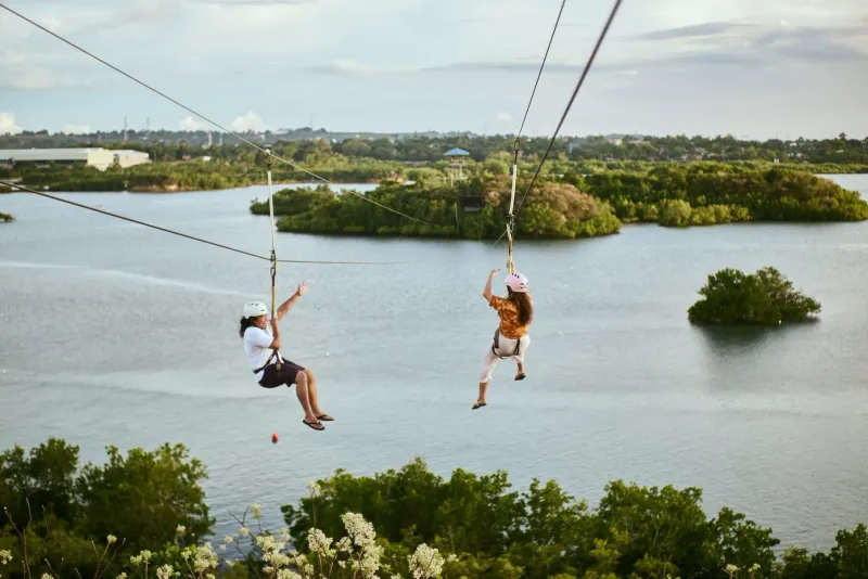 Two riders ziplining above calm water in a jungle adventure park similar to those on Koh Samui