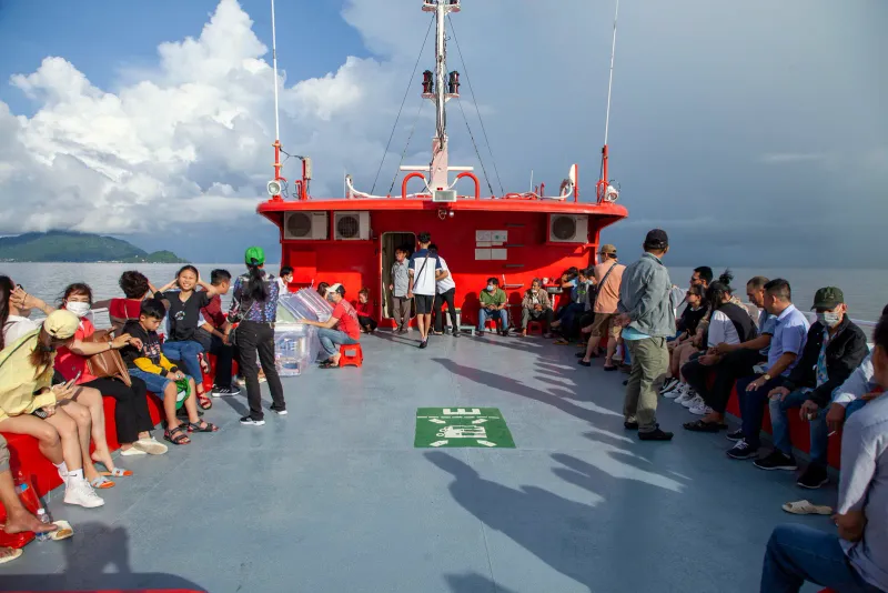 Passengers sitting on the open top deck of a red-bowed ferry with a green island on the horizon