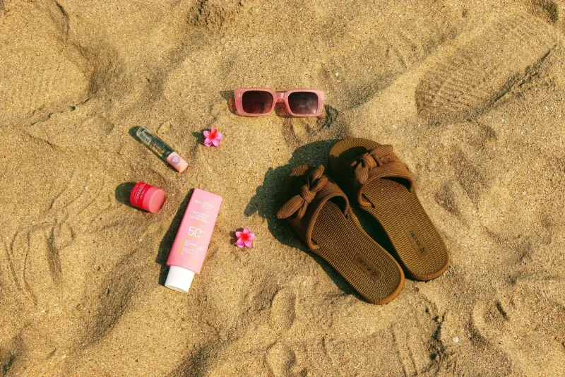 Flat lay of beach packing essentials for Koh Samui including sunglasses, sandals and reef-safe sunscreen on sand