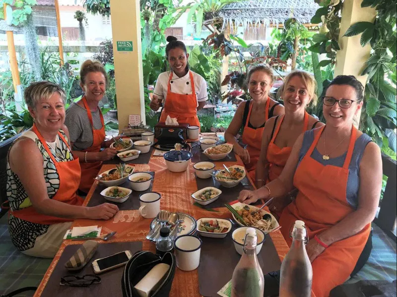 Guests in orange aprons smiling at a table of Thai dishes with the teachers standing behind them