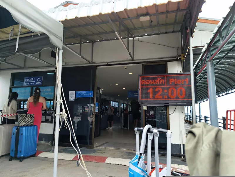 Ticket counters and a departure board at Nathon ferry pier on Koh Samui with travellers and suitcases