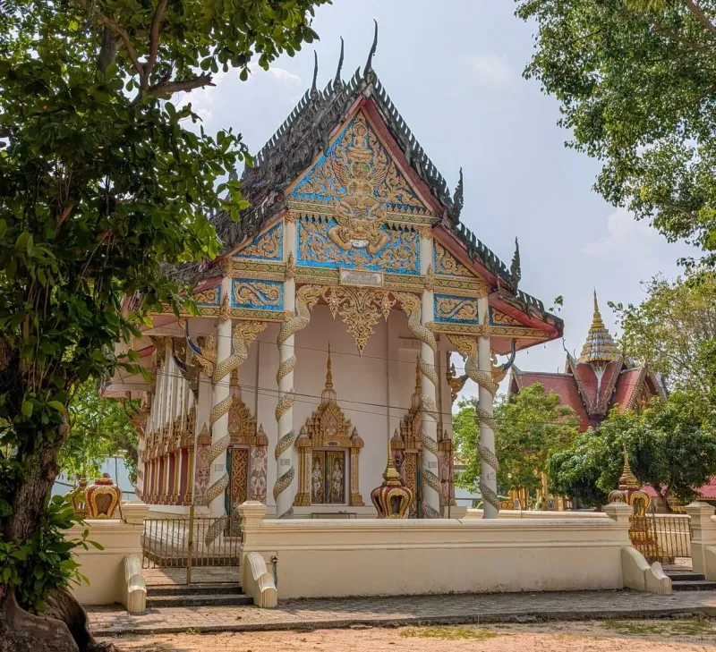 White and gold Thai ordination hall with naga-wrapped columns under shade trees
