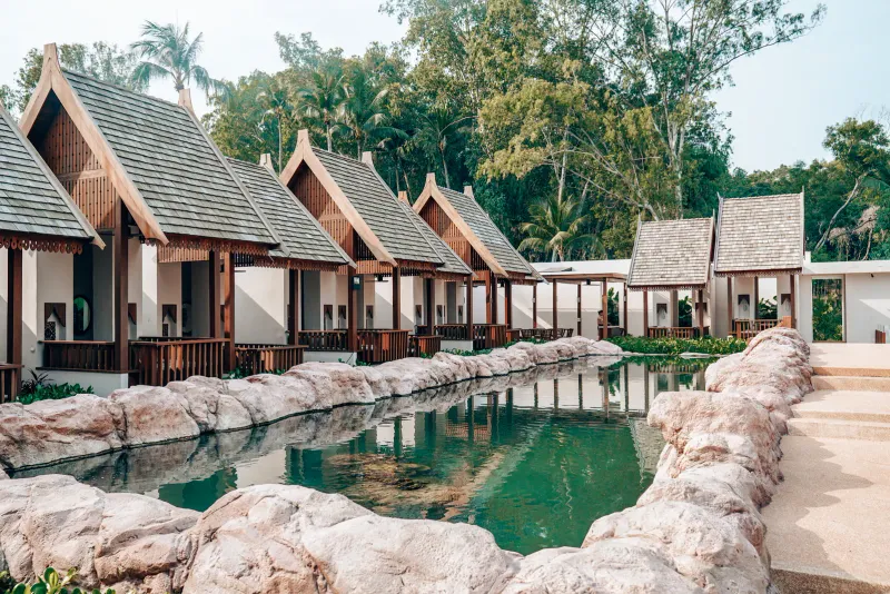 Row of Thai-style timber pavilions with pitched roofs along a rock-edged pond in the resort grounds