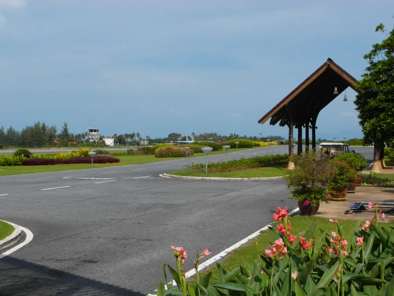 Open-air Samui International Airport apron with a timber roofed shelter and control tower in the distance