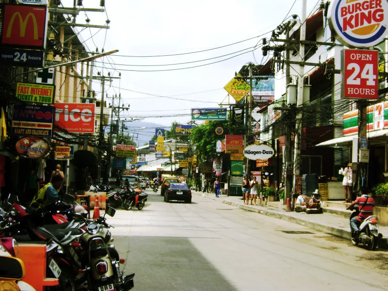 Busy Chaweng street with parked scooters, shop signs and a taxi in the distance on Koh Samui