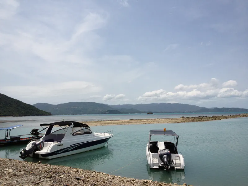 Two small speedboats moored in turquoise shallows off a rocky Koh Samui shore with hills behind