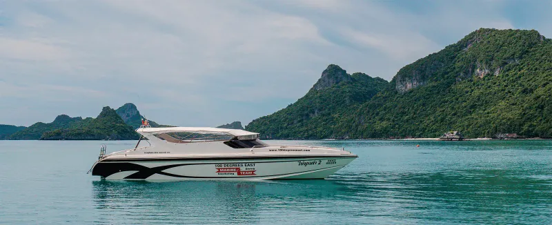 White speedboat anchored in calm green water beside forested limestone islands in the Gulf of Thailand
