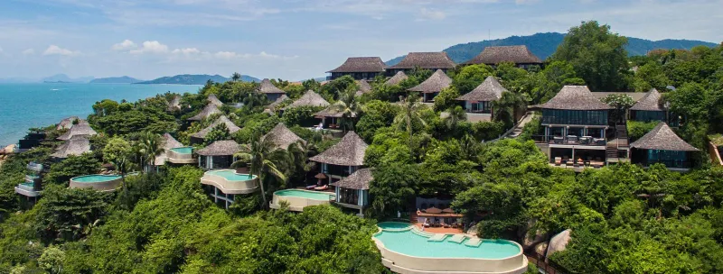 Aerial view of thatched-roof villas and stepped infinity pools on a forested hillside above the sea at Silavadee Pool Spa Resort