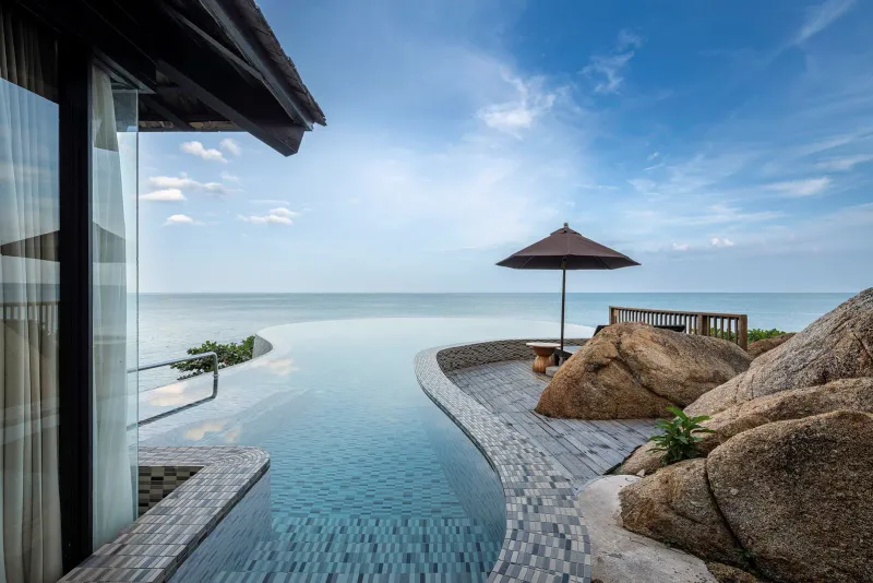 Curving infinity pool edge beside a villa, with granite boulders and open sea to the horizon