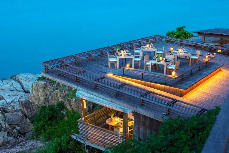 Clifftop dining deck at Six Senses Samui lit at dusk above the rocks and the sea
