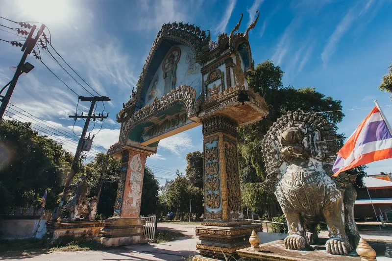 Ornate temple entrance archway painted with murals, flanked by a stone guardian lion and a Thai flag