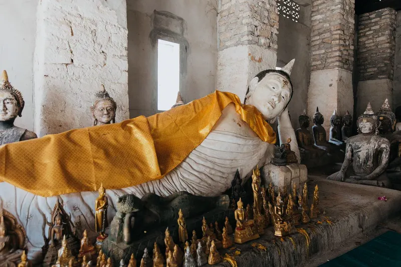 Reclining Buddha statue draped in a gold cloth surrounded by smaller votive statues inside a weathered stone hall