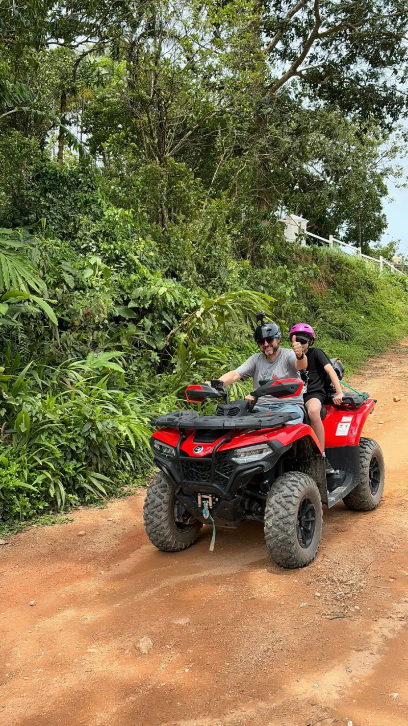 Two riders sharing a quad bike on a red dirt trail with a hillside white fence in the background, X-Quad Samui